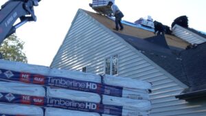 Stacks of plastic-wrapped GAF Timberline HDZ shingles in foreground, with roofers removing damaged shingles and underlayment from the peaked roof of a home with white lap siding